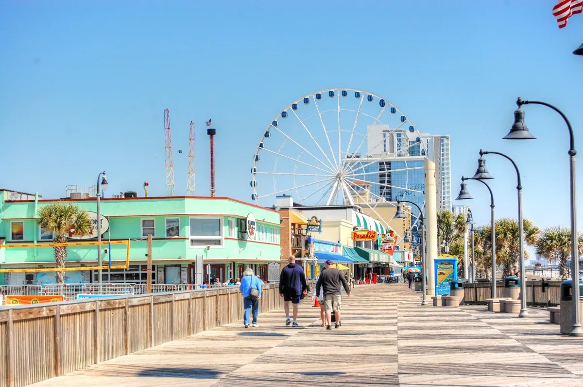 A vibrant exterior shot of the Myrtle Beach boardwalk, featuring the ferris wheel, colorful buildings, palm trees, and people strolling. The scene is lively and joyful, evoking the spirit of fun and exploration for all ages.