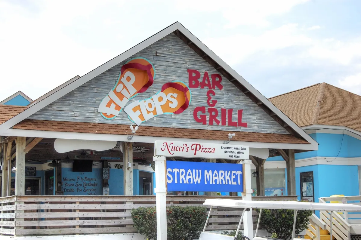 This exterior image focuses on signage for local eateries—Flip Flops Bar & Grill, Nucci's Pizza, and Straw Market—set against a backdrop of a relaxed, open-air environment. The scene hints at casual dining and socializing, perfect for unwinding after a day of adventure.