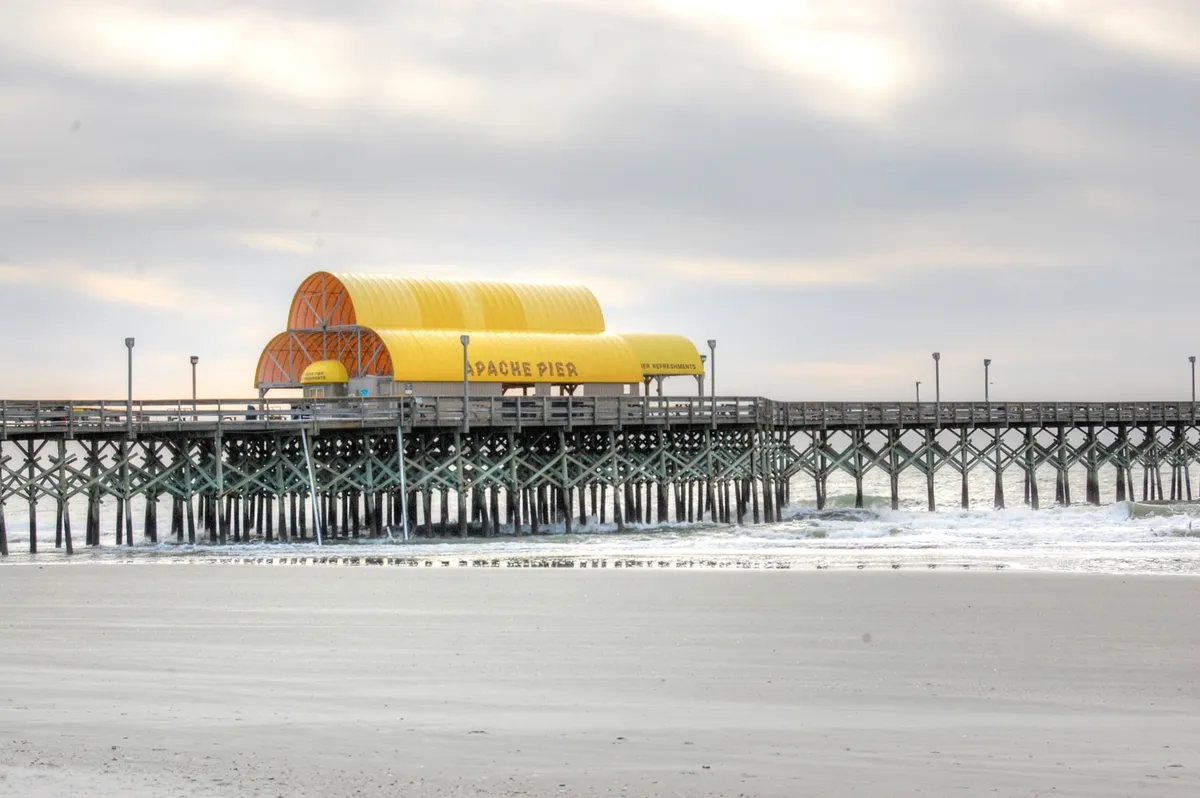 A wide, serene view of Apache Pier extending into the ocean, with gentle waves lapping below and a distinctive yellow roof structure providing a pop of color. The pier is surrounded by open sky and the distant coastline, inviting viewers to imagine relaxing walks or capturing beautiful coastal sunsets.