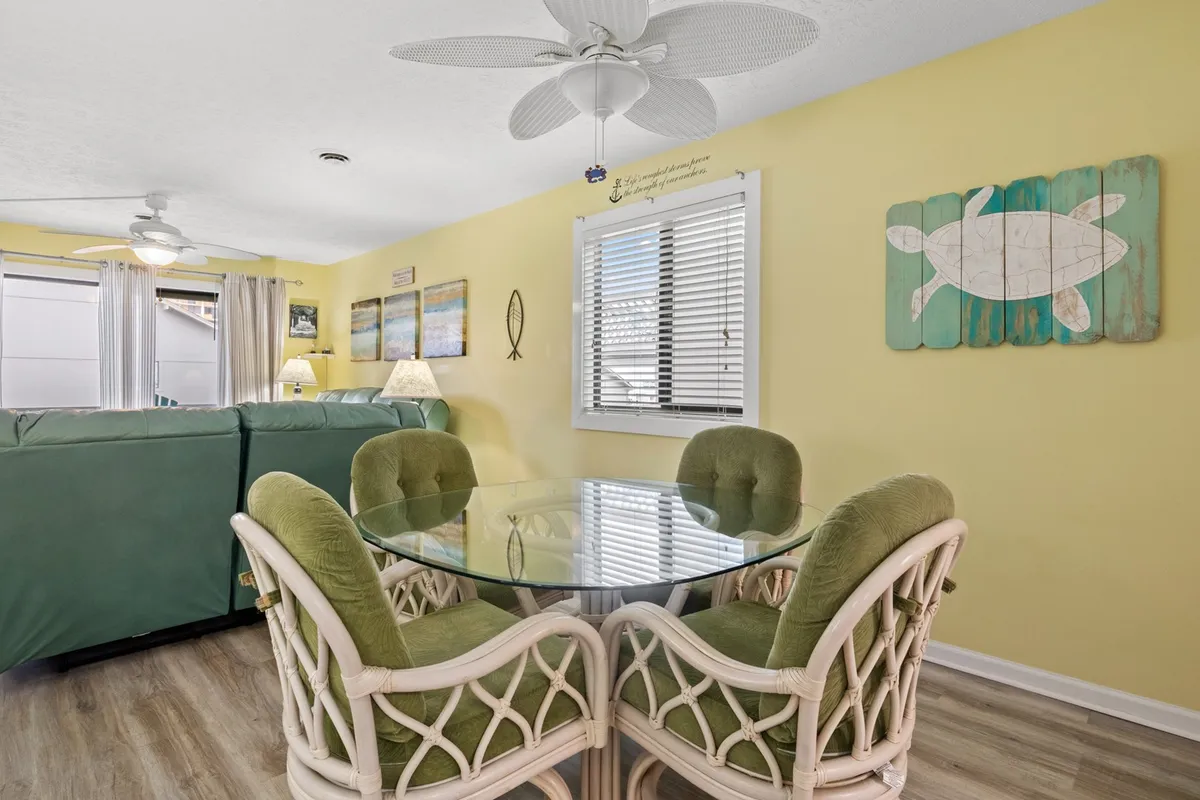 A bright dining area features a glass dining table with green upholstered chairs, turtle artwork, and side table lamps. The cheerful setting feels inviting and is suited for group meals or festive celebrations.