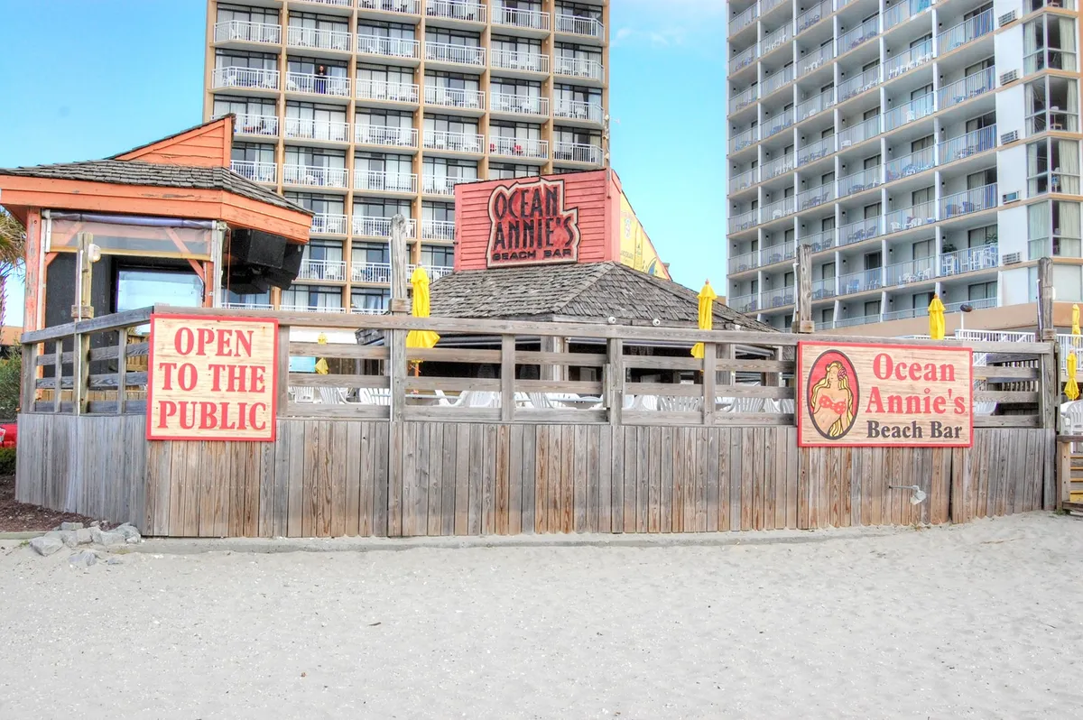 This vibrant exterior shot captures the entrance to Ocean Annie's Beach Bar, with its iconic signage, yellow umbrellas, and inviting, beachside atmosphere. The scene is lively and welcoming, suggesting fun and relaxation close to the condo.
