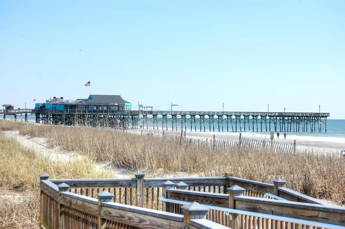 A breathtaking view of the beach featuring a wooden pier stretching into the blue Atlantic, soft dune grasses framing golden sand, and a bright sky overhead. People can be seen walking and enjoying the shoreline, with gentle waves rolling in.