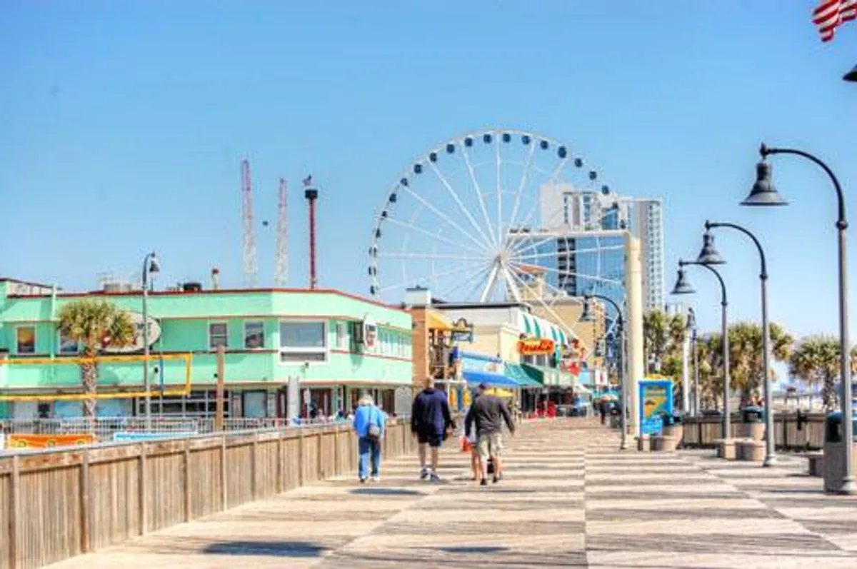 This lively exterior image captures Myrtle Beach's iconic boardwalk featuring a vibrant ferris wheel, colorful buildings, and visitors strolling along the path. The scene is bustling with activity, highlighting both the fun and variety available just minutes from the property.