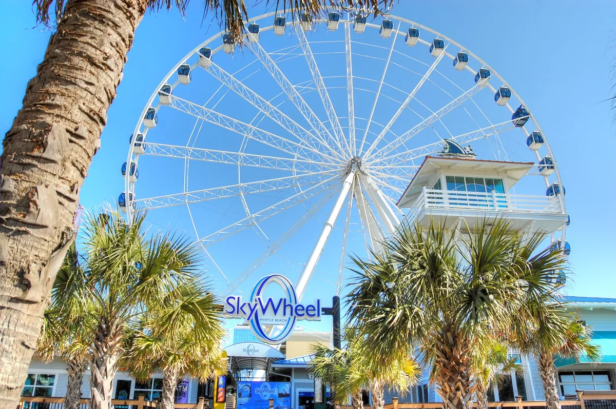An exterior scene featuring the SkyWheel—a major Myrtle Beach attraction—framed by palm trees and the entrance structure. Visitors can be seen gathering near the entrance, hinting at the excitement and adventure that awaits.