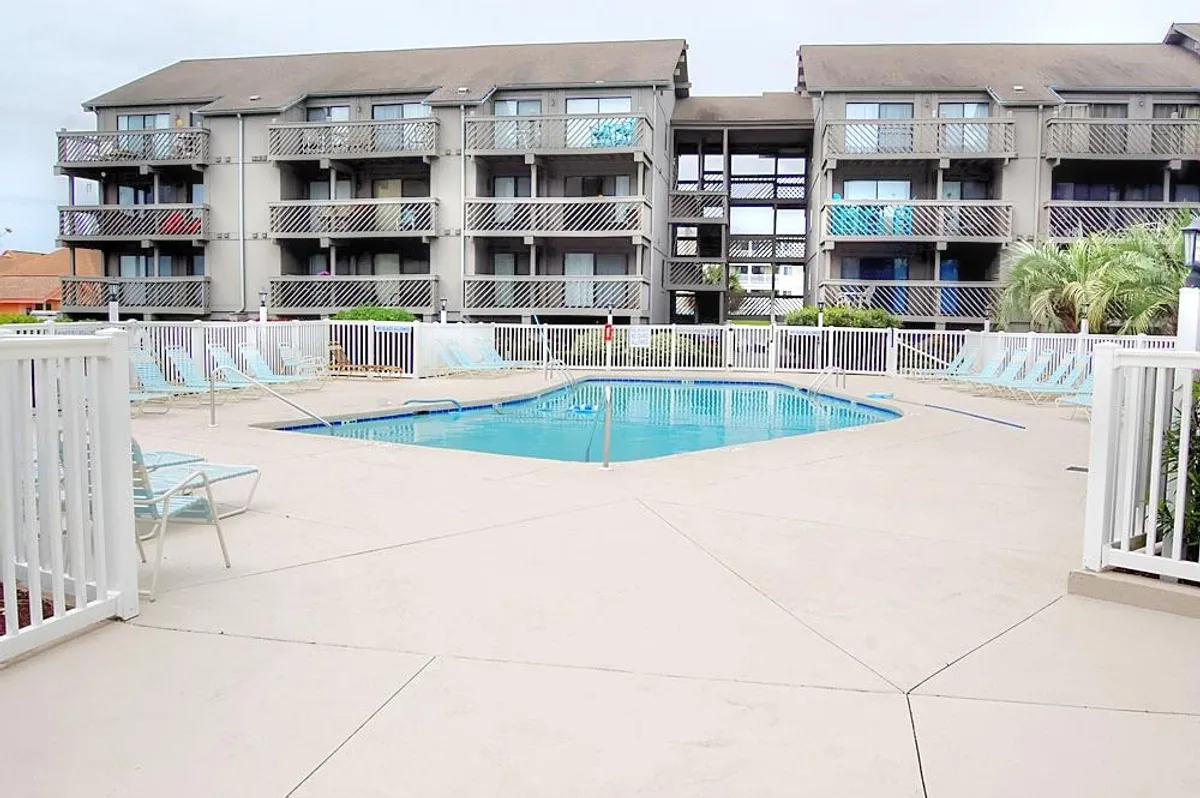 A sparkling outdoor swimming pool surrounded by ample lounge chairs, palm trees, and the balconies of Shipwatch Pointe I. The pool area is bathed in sunlight, with inviting clear water and a relaxed resort feel.