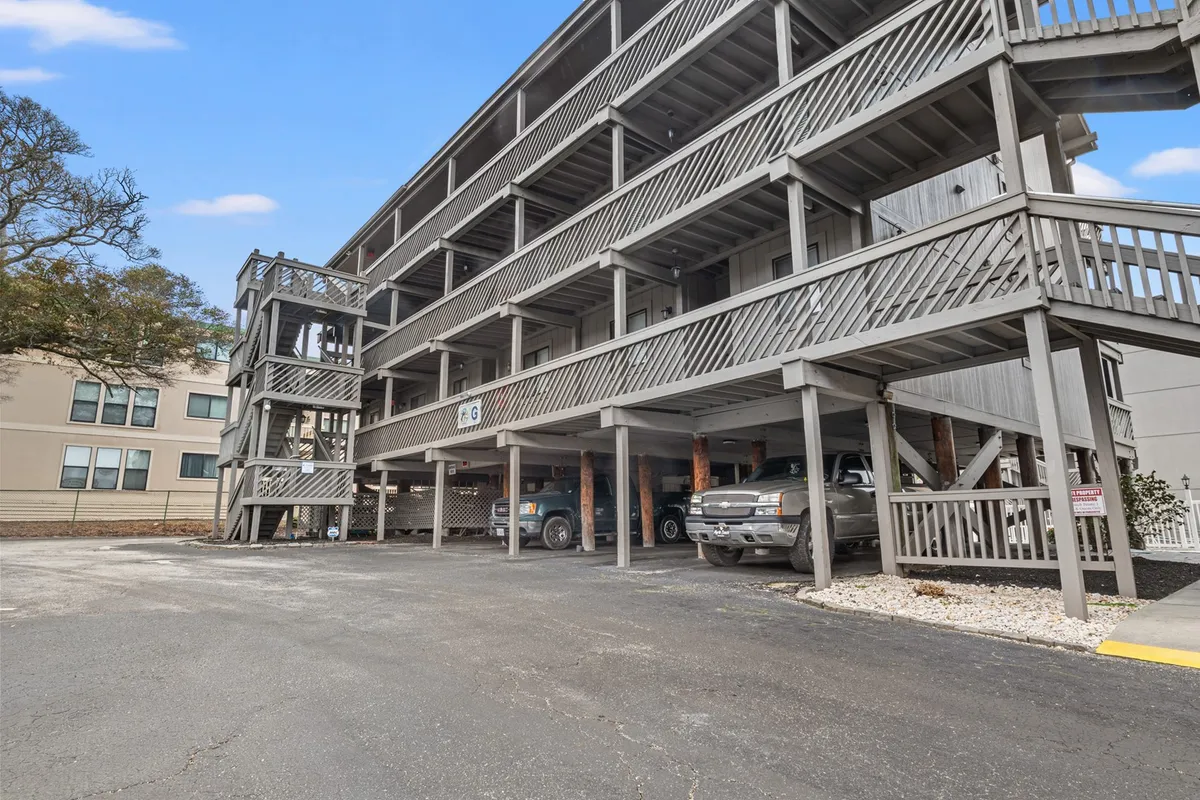 This exterior image shows the Shipwatch Pointe condo building with its distinctive wooden balconies, staircases, and parking area. The scene hints at the communal outdoor spaces where guests might gather, relax, or enjoy the sunset from their balconies.