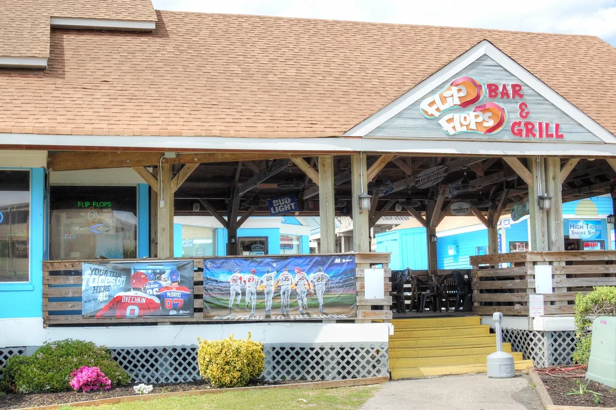 This image features the colorful signage and outdoor seating area of 'Flip Flops Bar & Grill,' with sports-themed banners and a lively patio setup. The casual, cheerful mood encourages social activities and outdoor dining.