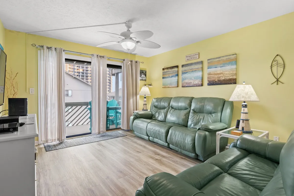 The hero image showcases a bright, cozy living area with plush green leather sofas arranged for socializing or relaxing, unique coastal wall art, a charming lighthouse lamp, and a balcony door letting in an abundance of natural light. The inviting setup hints at leisurely afternoons indoors and easy access to the balcony for a breath of fresh sea air.