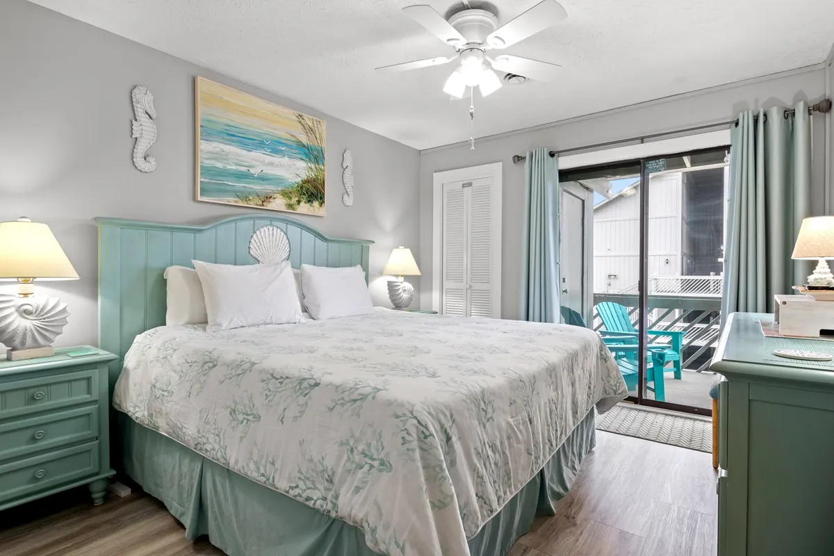 A tranquil bedroom featuring a decorative headboard on the bed, a painting of a coastal scene above, and soft light streaming in from a window. The room includes elegant light fixtures and a glimpse of a private balcony, suggesting opportunities for relaxation and ocean views.