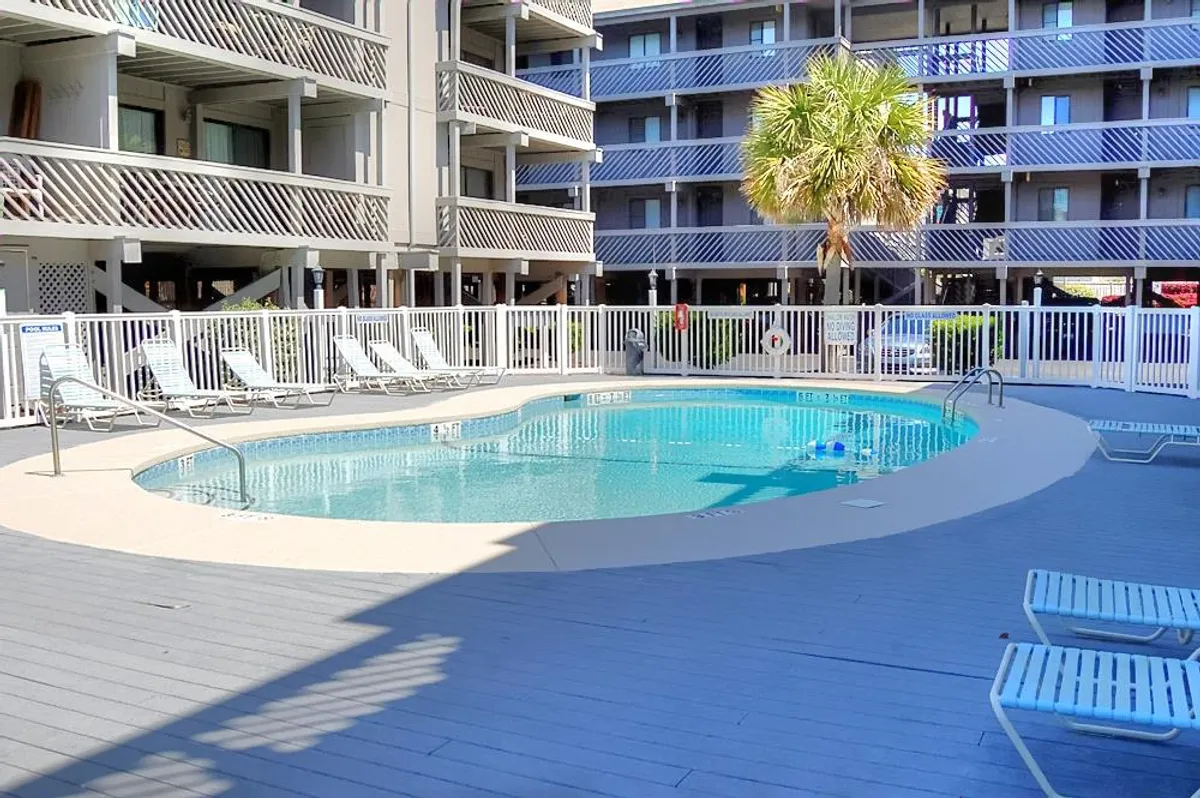 Another perspective of a large swimming pool with comfortable lounge chairs, a prominent palm tree, and the building facades in the background. The scene is active and social, ideal for groups or families.