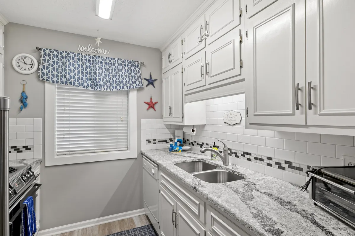 A bright, inviting kitchen featuring a spacious granite countertop with a double sink. The window above is dressed with cheerful curtains, and whimsical decorative stars and a clock add homey touches. The generous counter space and layout suggest plenty of room for meal prep and family gatherings.