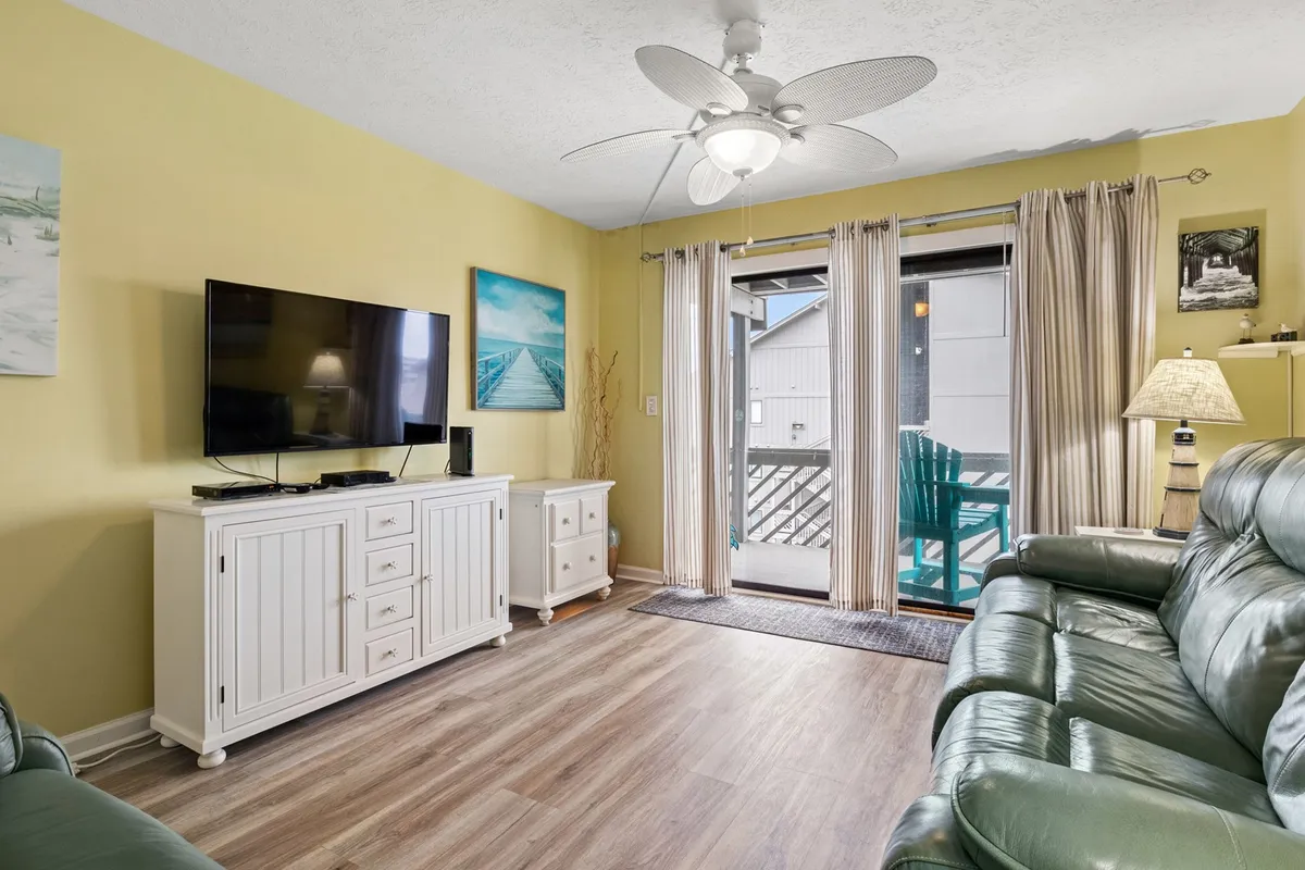 The image showcases a spacious open-concept living area with a comfortable sofa, TV, decorative art on the walls, and a sliding glass door that leads to a balcony. Natural light fills the room, creating a bright and welcoming atmosphere ideal for relaxation or social gatherings.