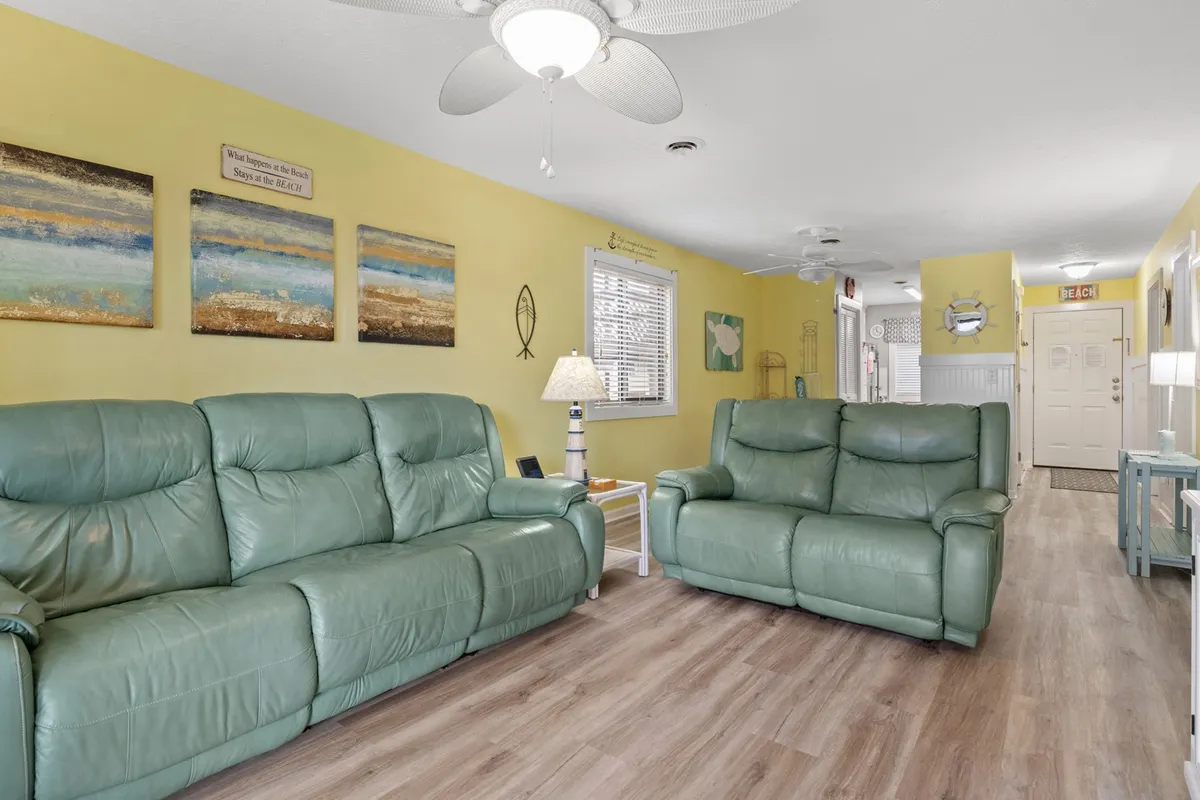 A bright, inviting living room featuring green leather sofas arranged for conversation, ocean-themed wall art, and a sunlit window. The cheerful, cozy space suggests a comfortable home base for extended winter stays, social gatherings, or simply relaxing after exploring the area.